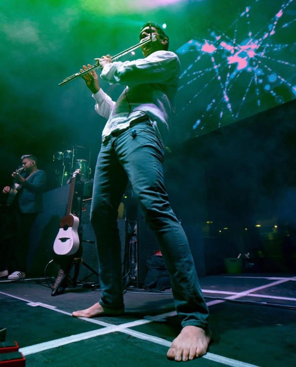 Huáscar Barradas tocando descalzo en un escenario grande, con su banda detrás y las luces de fondo.