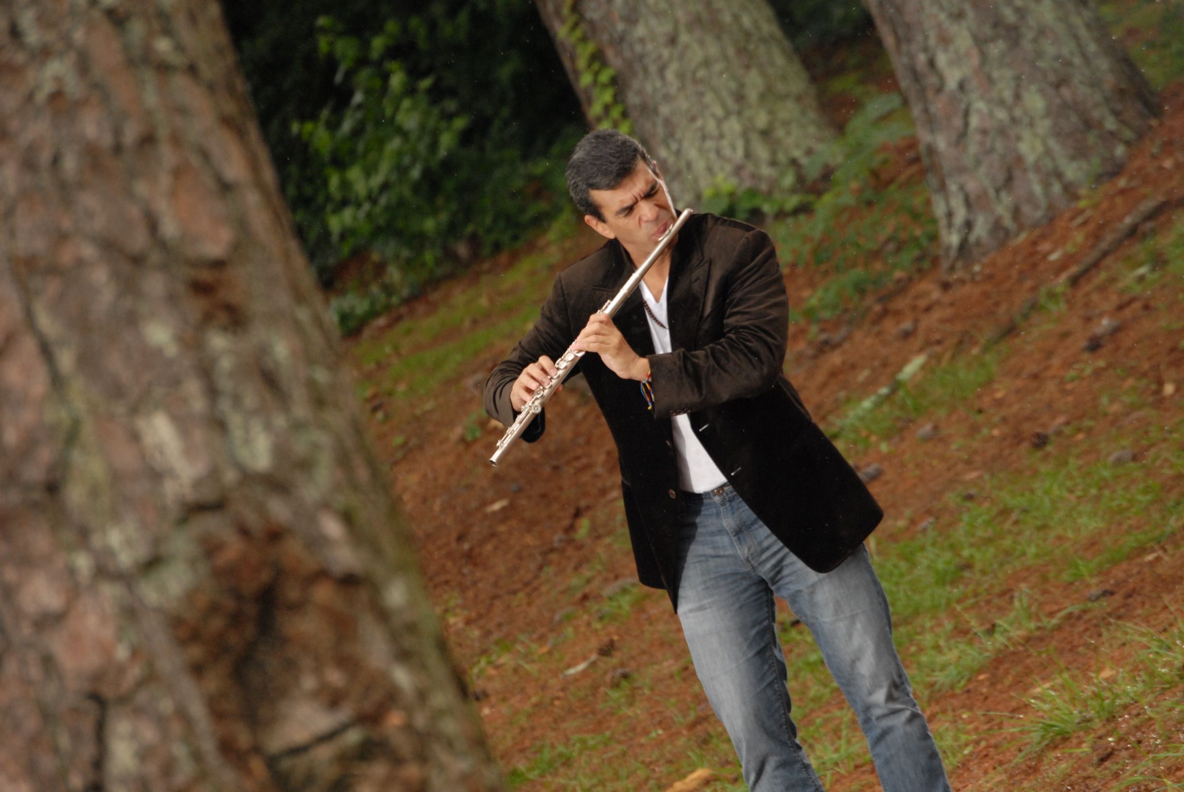 Huáscar Barradas tocando la flauta de pie en un bosque de pinos, con saco oscuro.