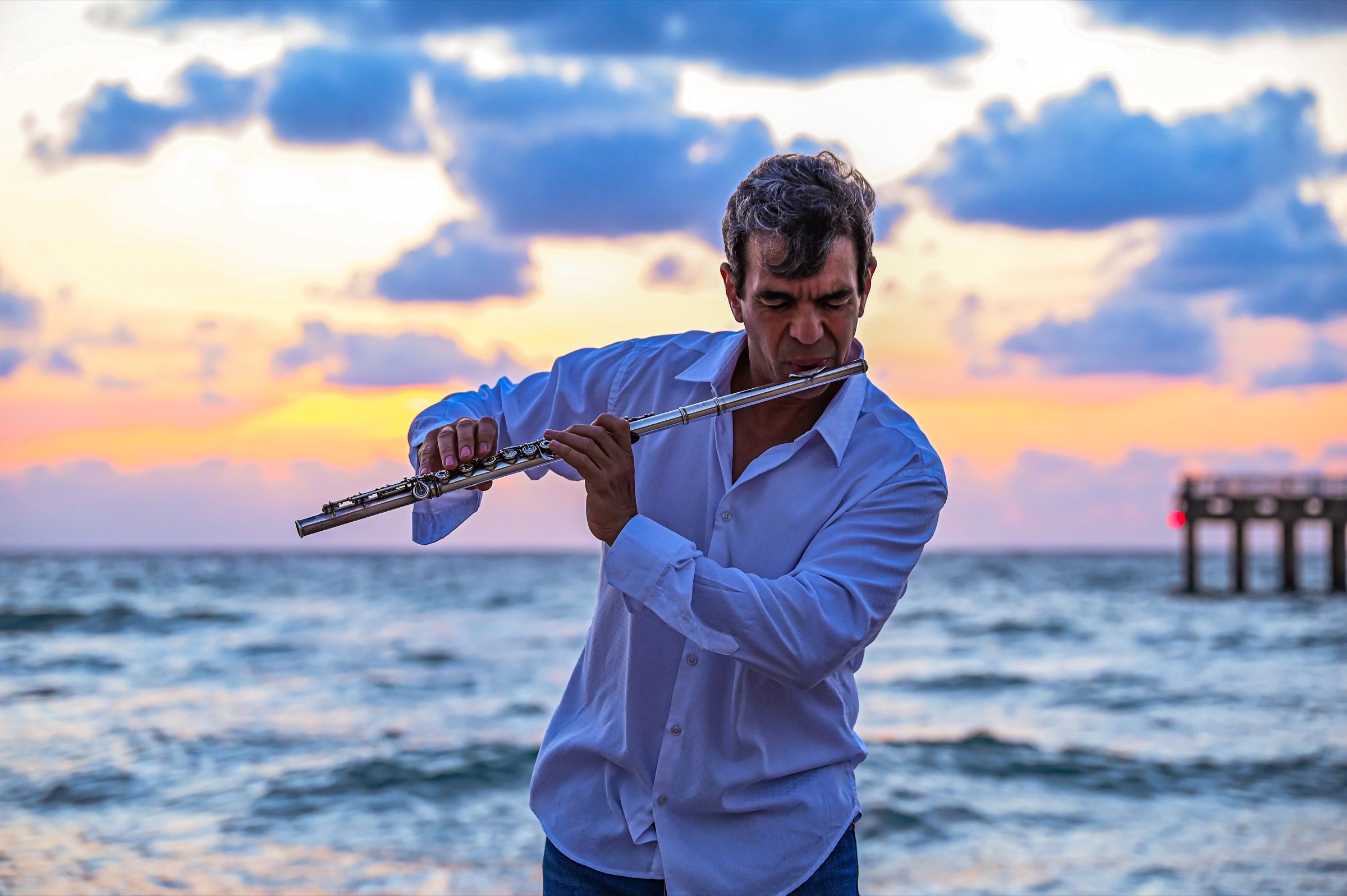 Huáscar Barradas tocando la flauta a la orilla del mar, con el cielo del atardecer detrás.