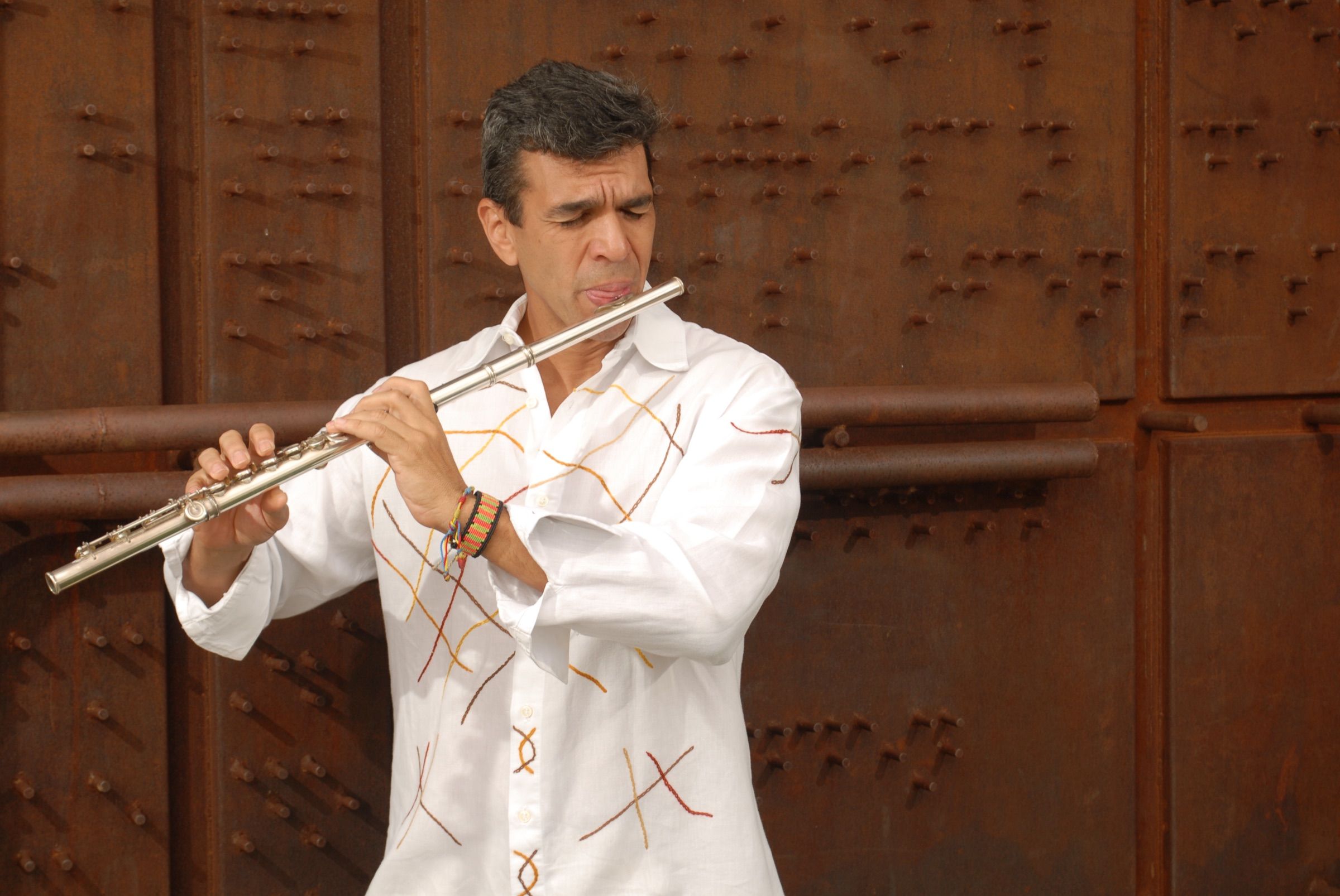 Huáscar Barradas tocando la flauta con camisa blanca, frente a un portón de acero oxidado.