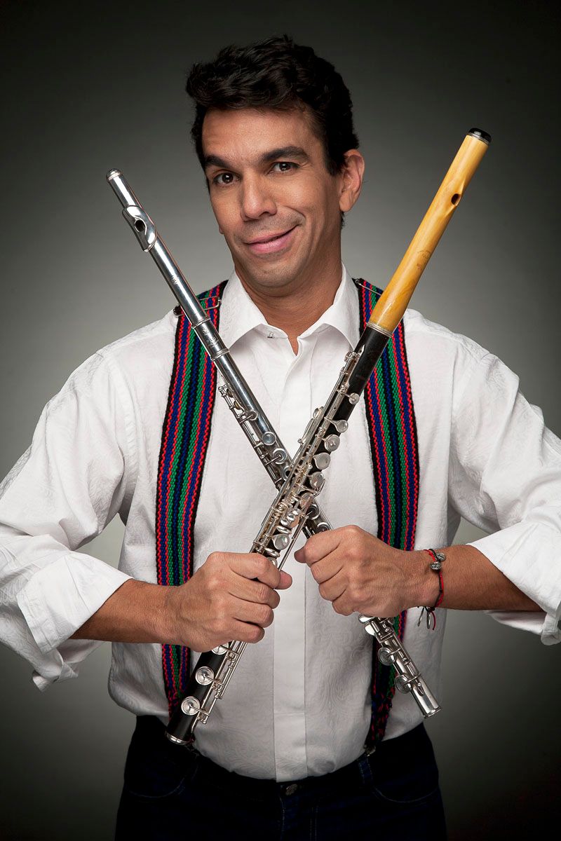 Huáscar Barradas de camisa blanca y tirantes de colores, sonriendo a cámara con dos flautas cruzadas.